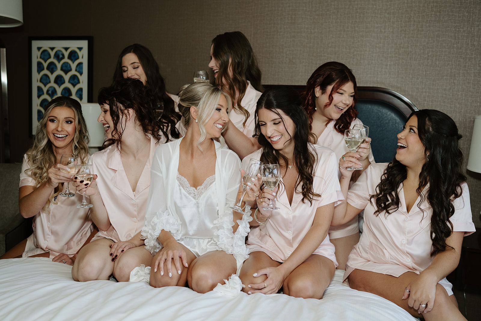 Bride and bridesmaids laugh together while holding wine glasses during wedding morning preparations.